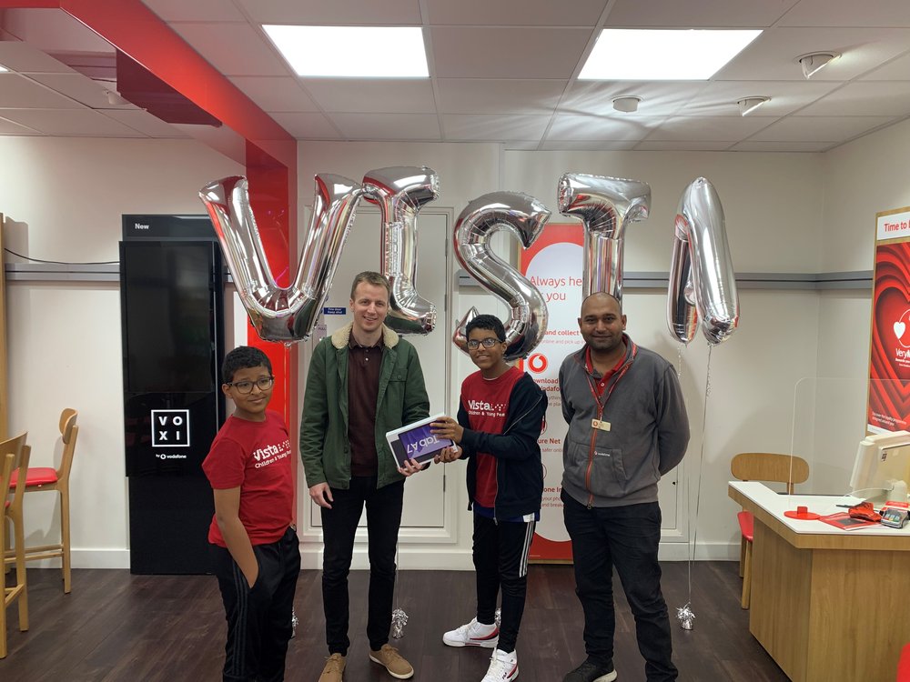 Image is of staff at the Vodafone store n Loughborough standing with the winner of Vista's virtual balloon race holding the tablet in front of balloons that spell out 'Vista'.